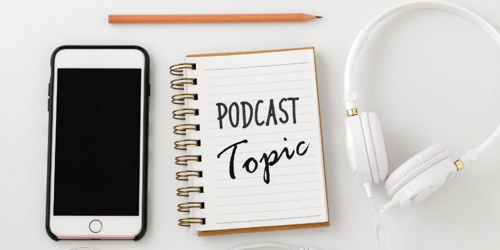Overhead view of spiral notebook with 'Podcast Topic' written on it, next to smartphone and white headphones on clean desk