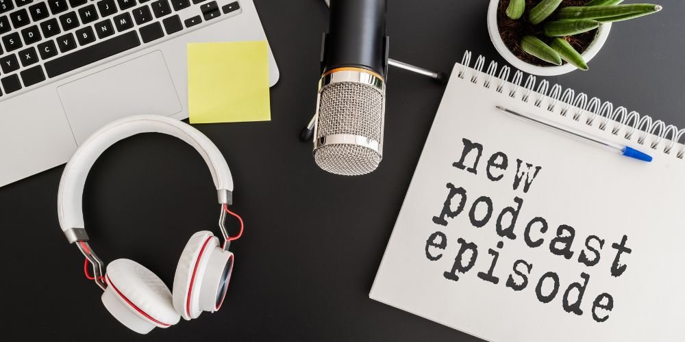 Overhead view of desk with notebook showing 'new podcast episode' text, professional microphone, headphones, and laptop for podcast creation